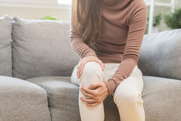 Health care of knee pain concept, close up hand of woman arthritis sitting joint ache, sore cramp or sprain tendon in leg on sofa at home. Osteoarthritis chronic disease of body problem from trauma.